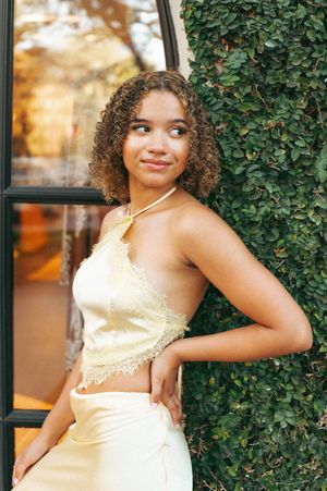 Young woman in yellow crop top and skirt posing outdoors against green ivy wall near window.