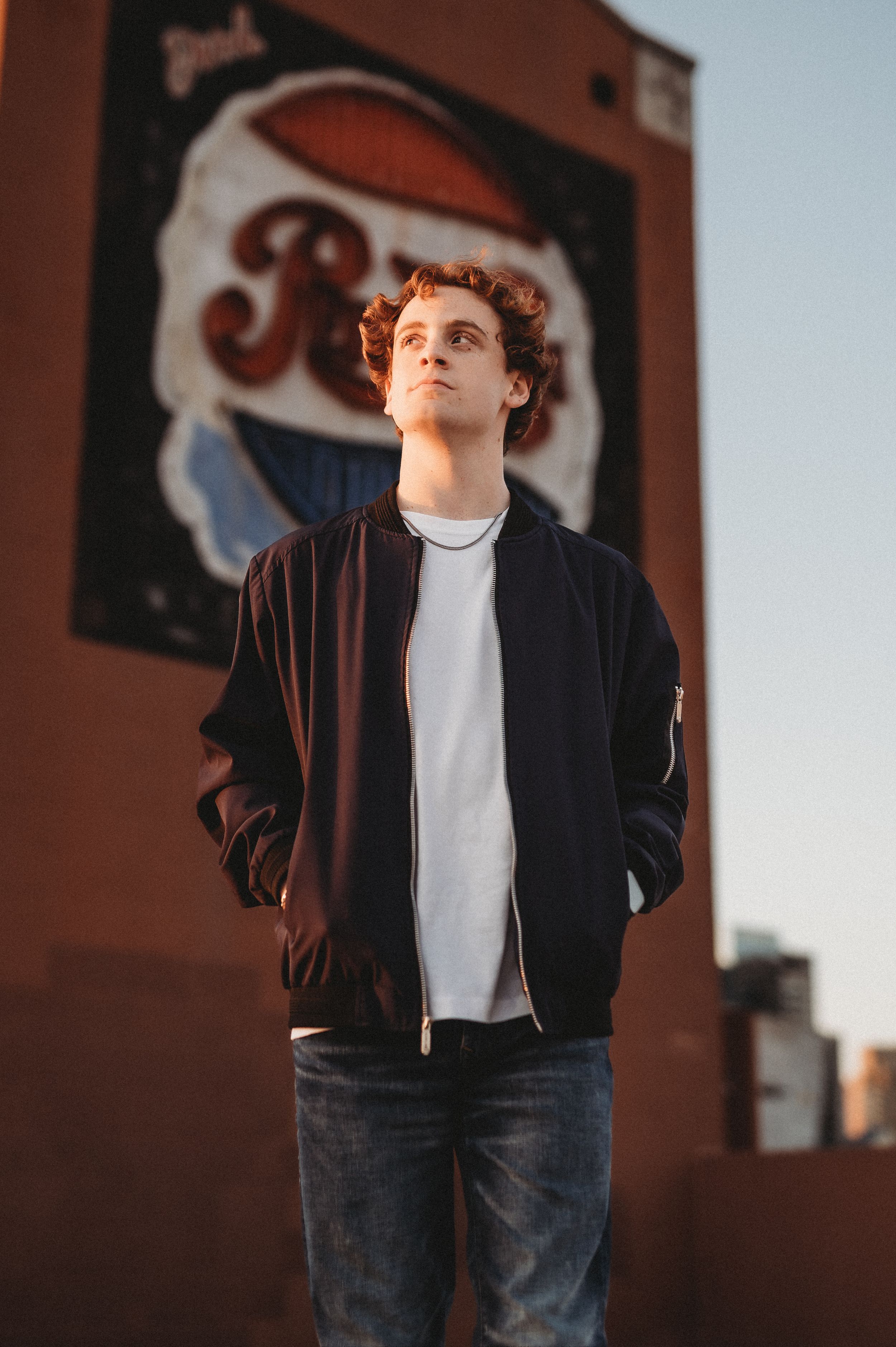 Person in brown bomber jacket and jeans standing against urban wall with vintage signage in golden hour lighting.
