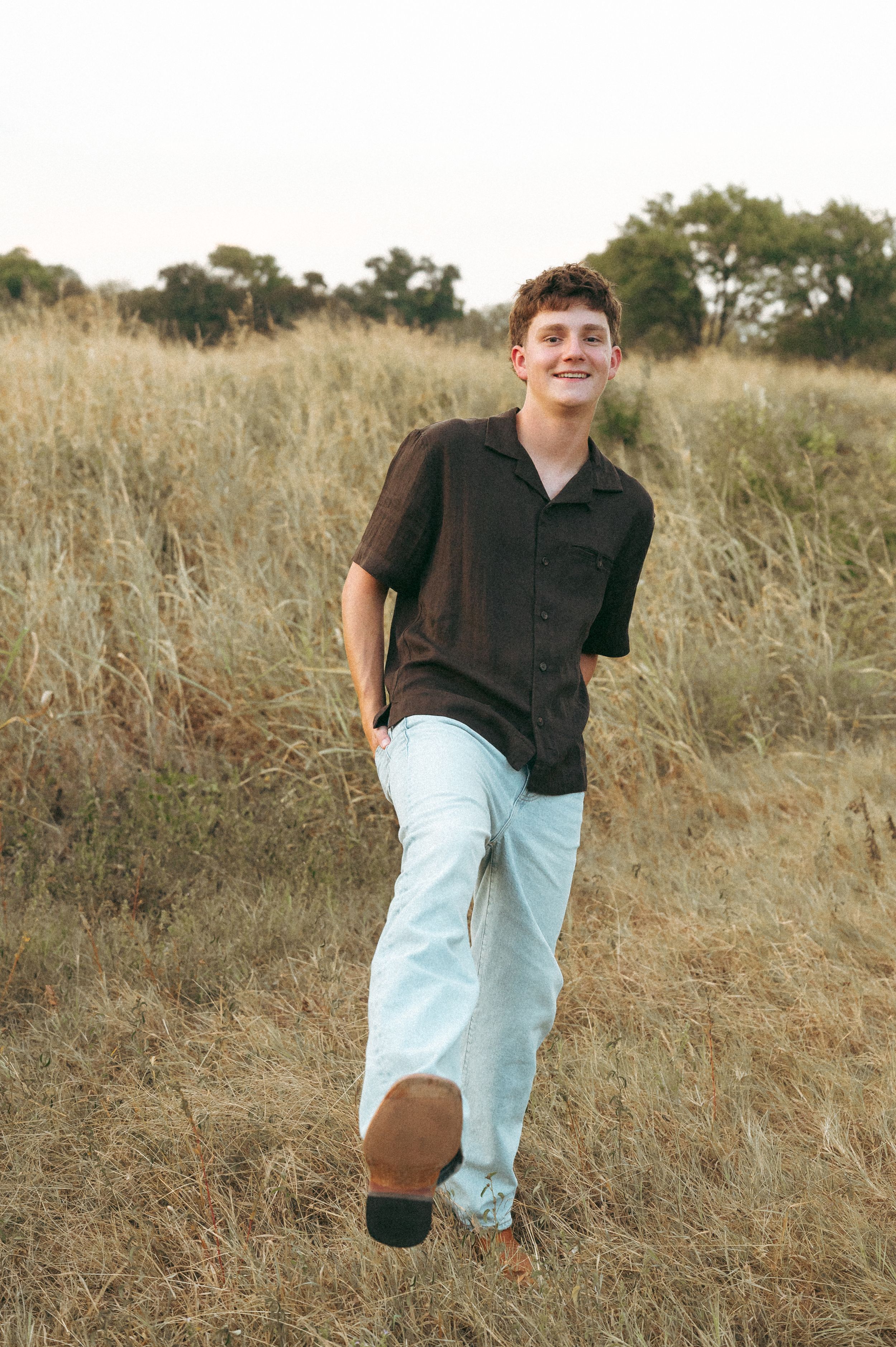 Smiling young man in brown shirt and light jeans walking through golden grass field with trees in background.