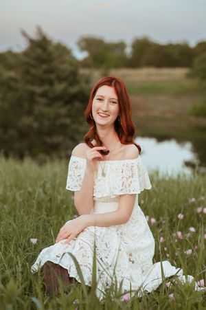 Young woman with red hair in white lace off-shoulder dress sitting in green field with wildflowers near pond.