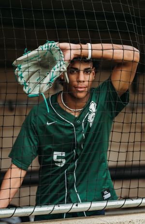 Baseball player in green Michigan State jersey stands in dugout behind protective netting.