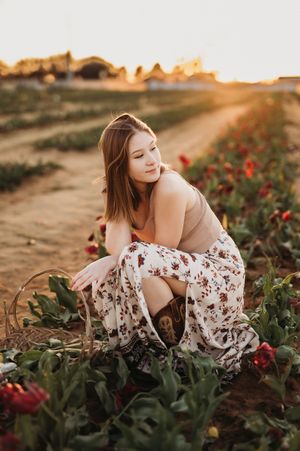 Person in floral skirt poses gracefully among red flowers on hillside during golden sunset hour.