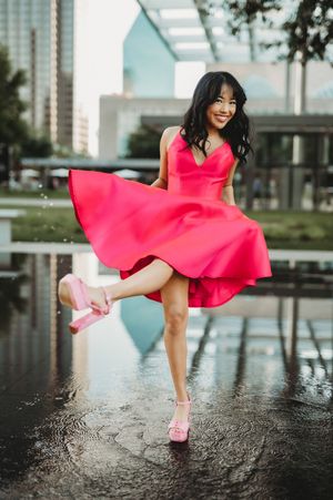 A person in a vibrant pink dress and matching heels twirls playfully near a reflective pool in an urban setting.