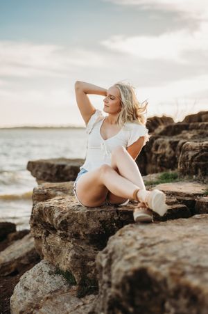 A person in a white dress poses on rocky coastal cliffs at sunset, with ocean waves and cloudy skies in the background.