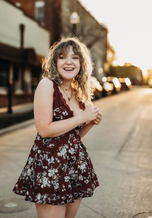 A person in a burgundy floral dress stands laughing on a sunset-lit street with storefronts in the background.