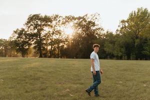Young man in white t-shirt and jeans walking in a sunlit green field with trees at golden hour.