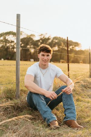 Young man in white t-shirt and jeans sitting by barbed wire fence in golden hour sunlight on a rural farm.