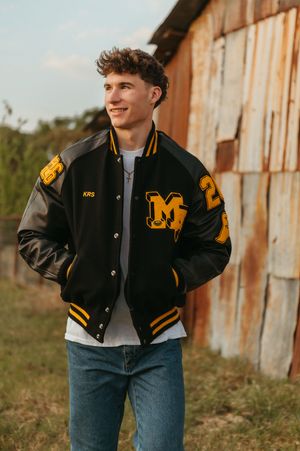Young man in black and gold varsity letterman jacket with M patch poses outdoors near rustic wooden wall.