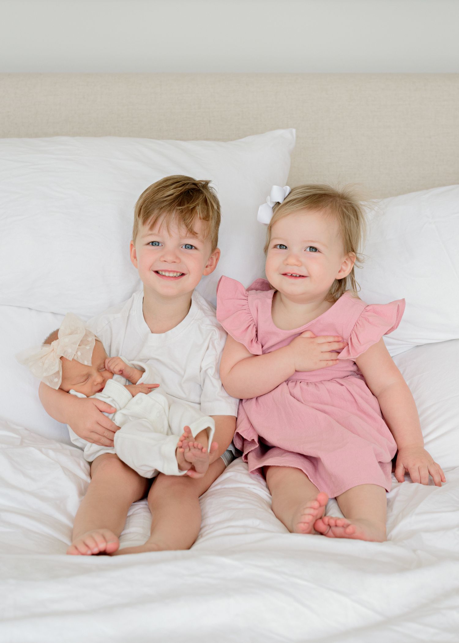 Two young children sitting on white bedding, wearing white and pink outfits while one holds a baby doll.
