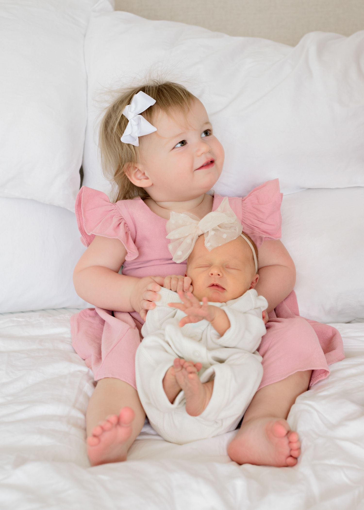 Young child in pink dress holds baby doll while sitting on white bedding with bow hair accessory.