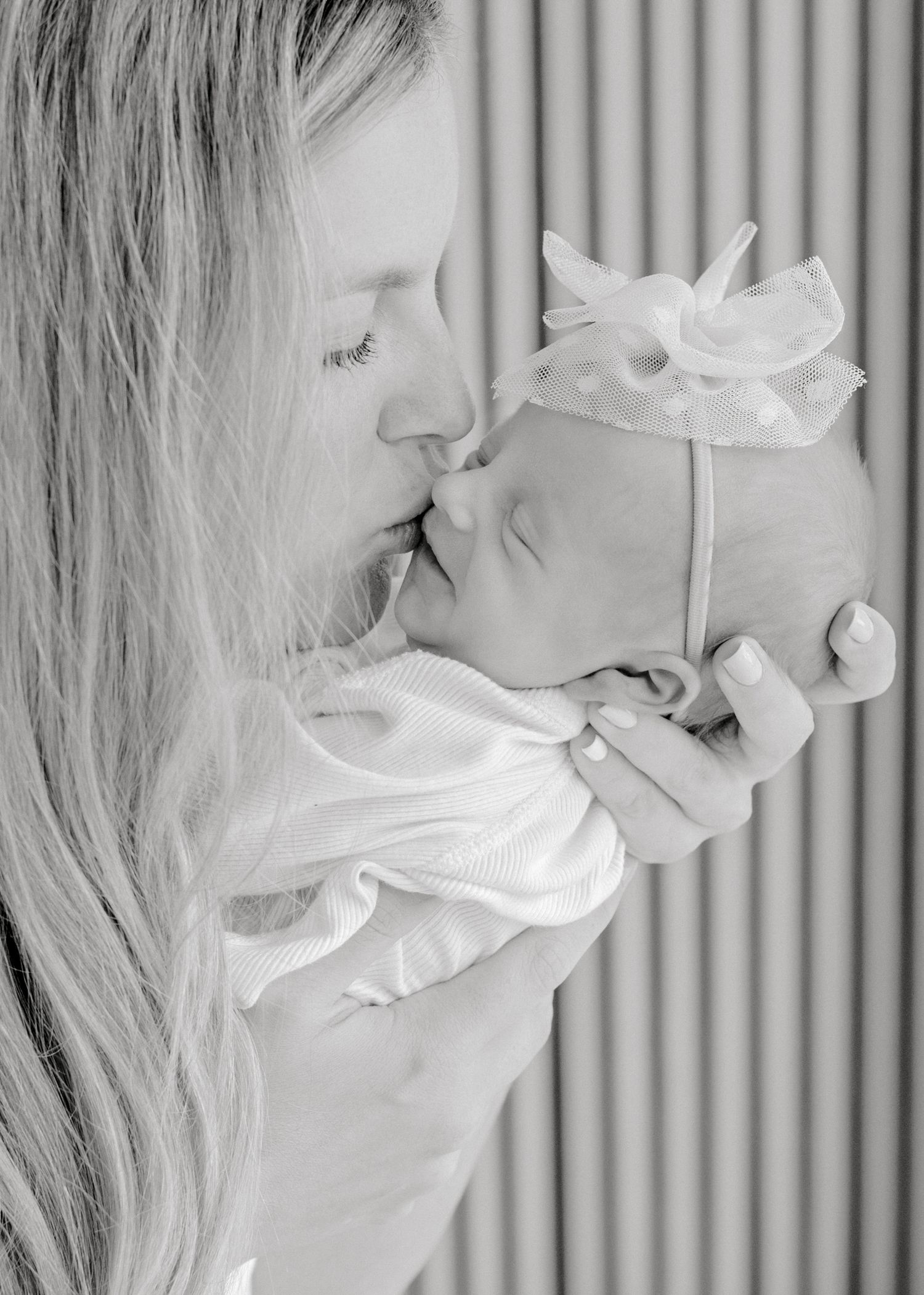 A tender black and white portrait of a parent and infant sharing a gentle kiss, with soft lighting and vertical stripes in background.
