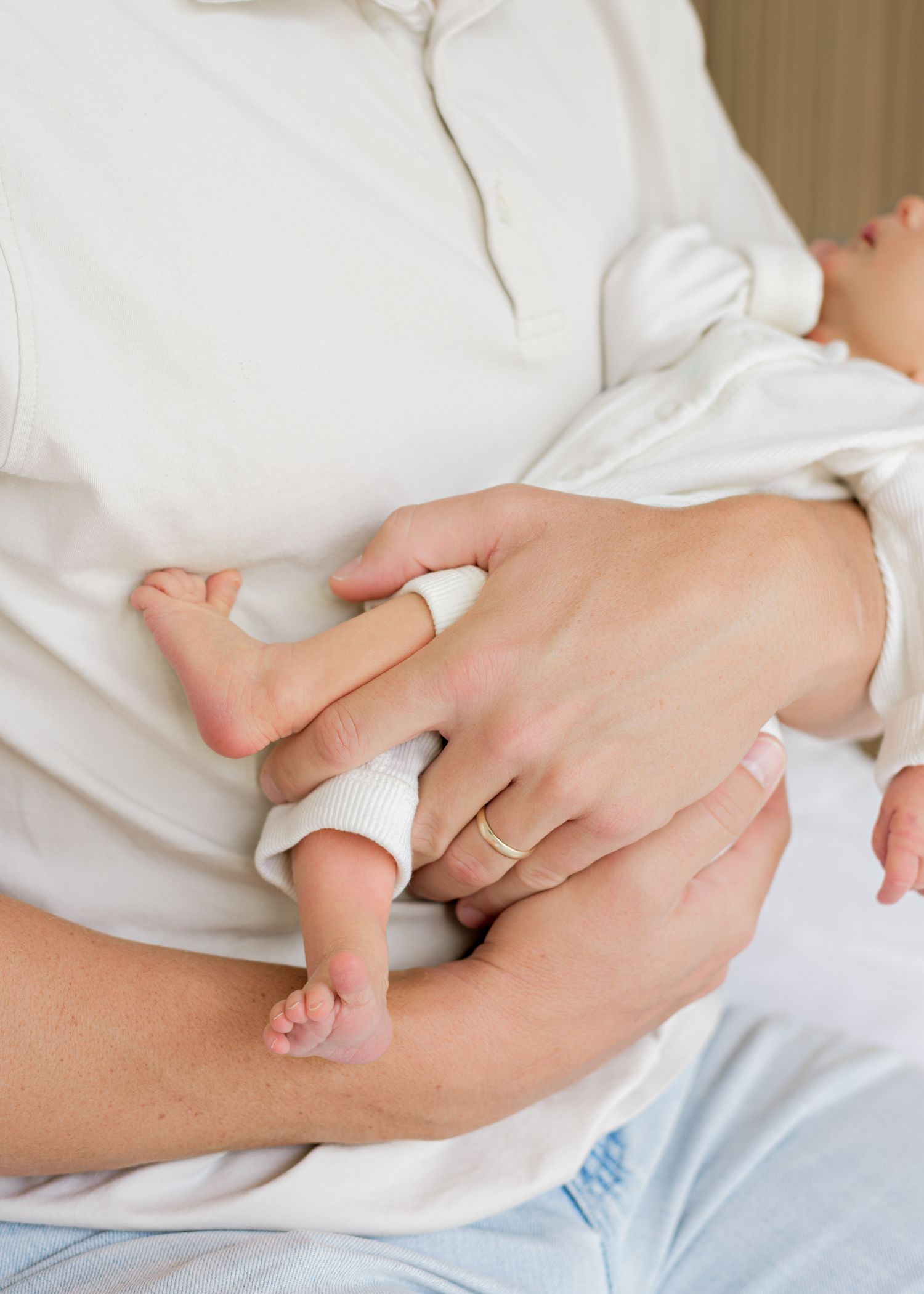 A parent's hands gently holding their baby's tiny feet while both wear white clothing on a bed.