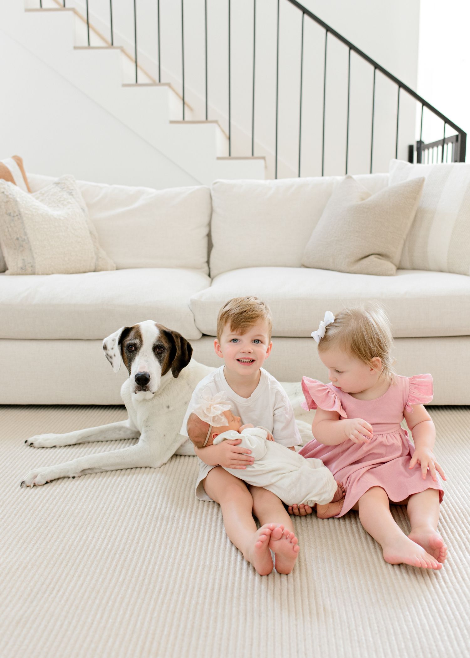 Two young children in white and pink outfits sit on a gray carpet with a spotted dog by a white couch and staircase.