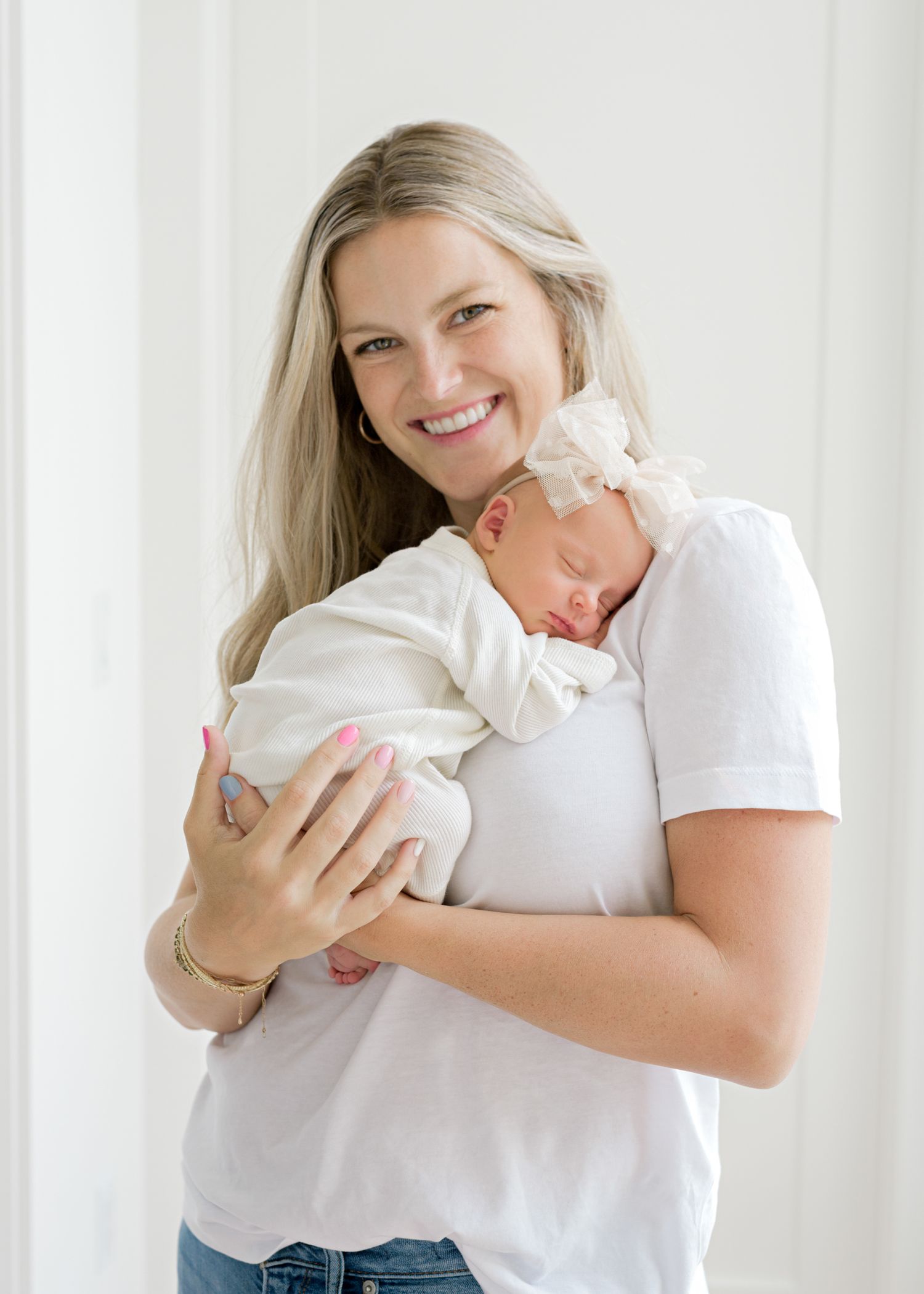 A tender moment captured as a mother in white t-shirt and jeans holds her sleeping newborn baby against her chest.