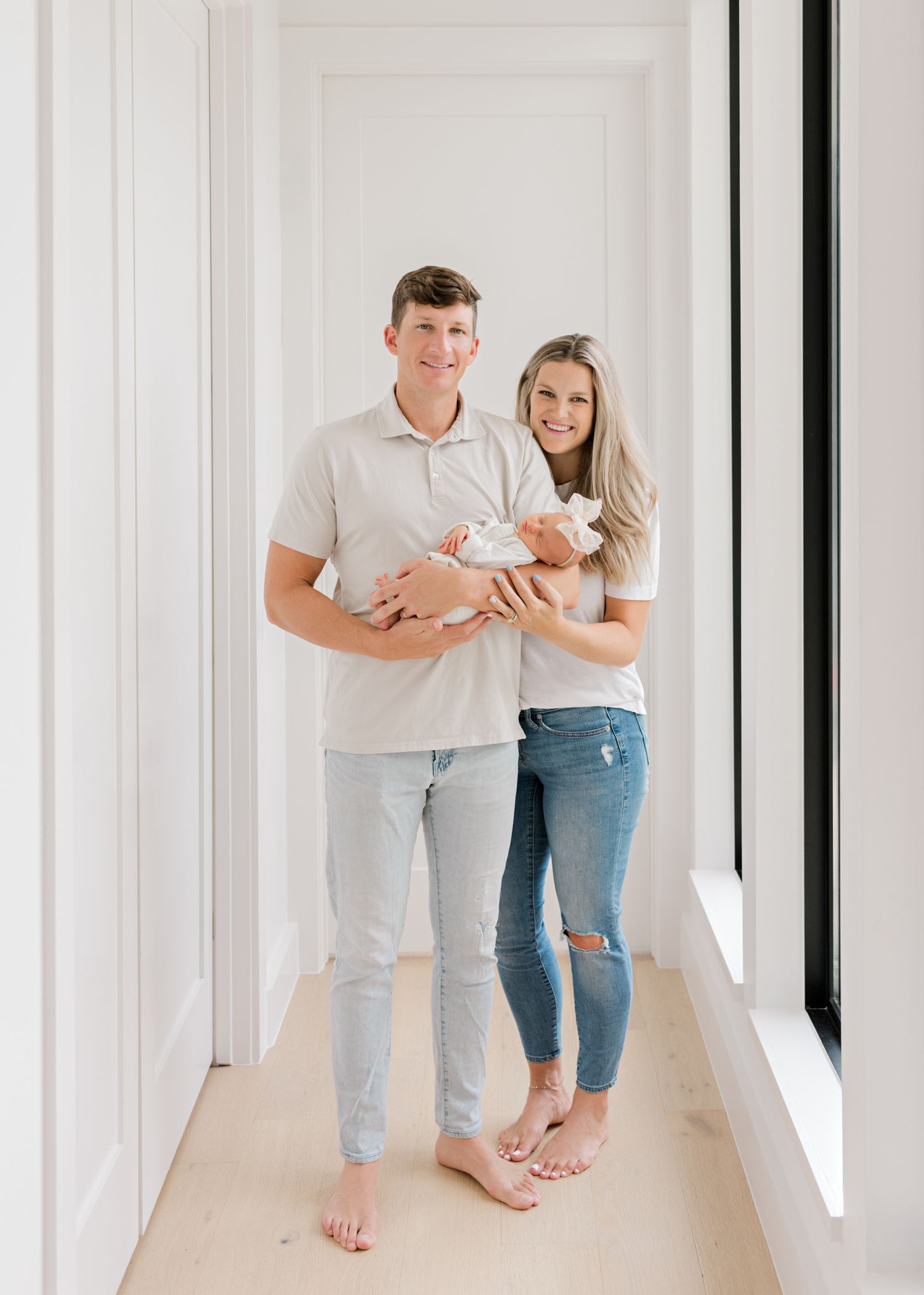 A couple stands barefoot in a bright white hallway while holding a newborn baby wrapped in blankets.