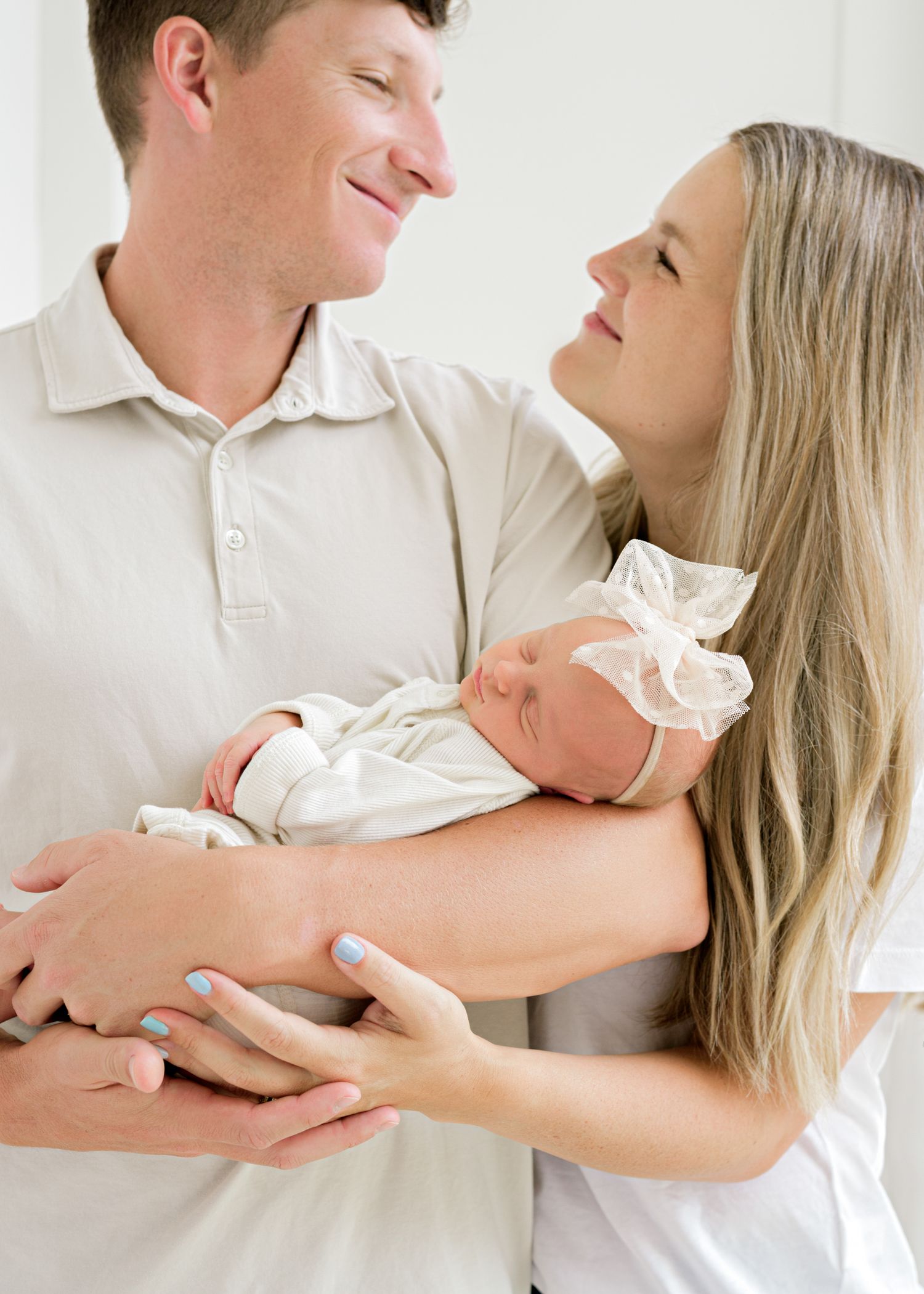 Happy couple dressed in white smiles lovingly at each other while holding their newborn baby in a tender family moment.