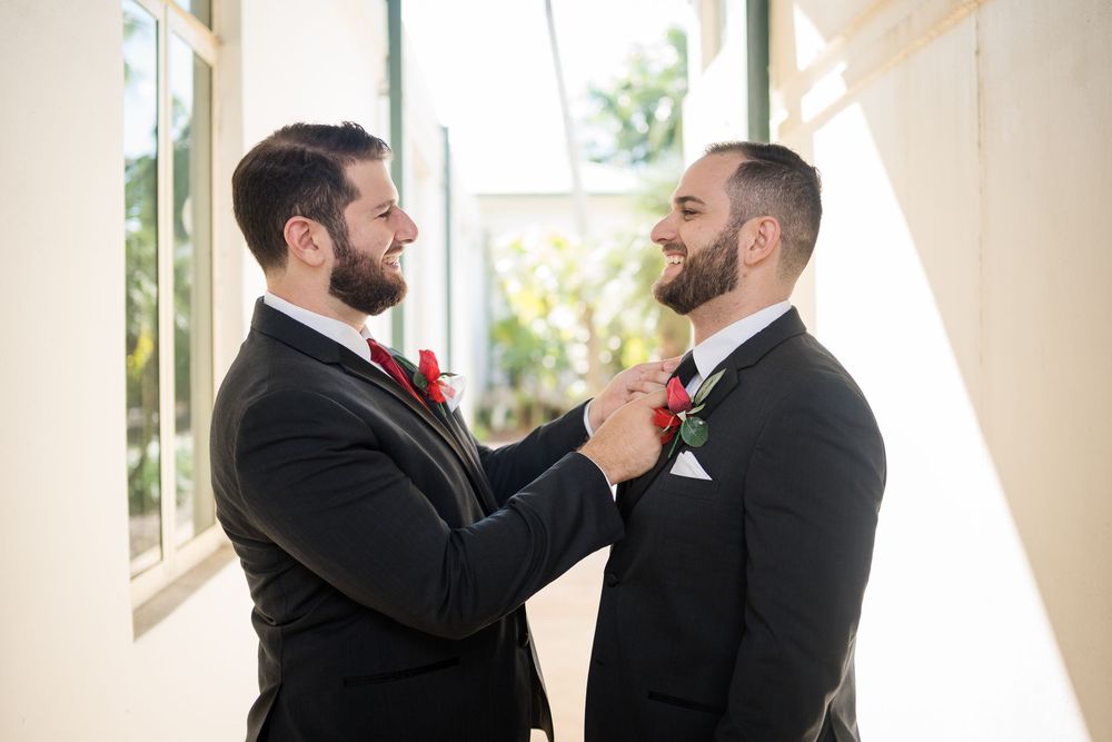 Two men in black suits adjust boutonnières while sharing a joyful moment in a sunlit hallway.