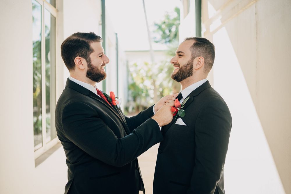 Two men in formal attire help each other with red floral boutonnieres during a wedding preparation.
