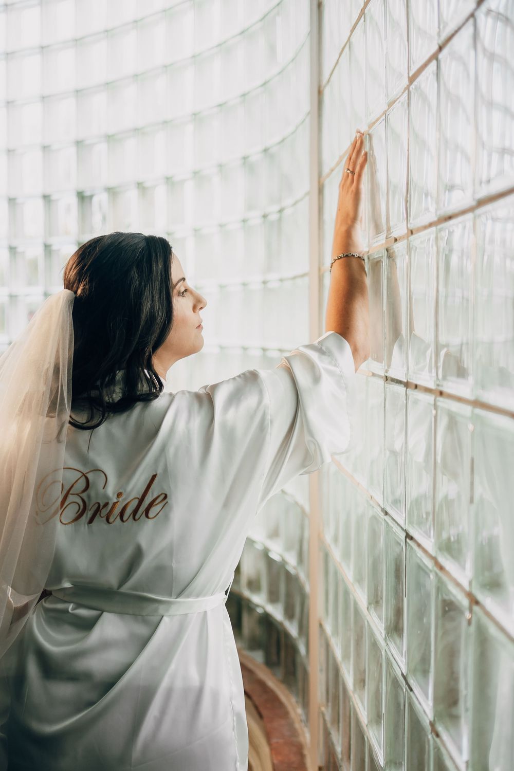 Someone in a white satin robe with 'Bride' embroidery reaches toward curtains in a light-filled room.
