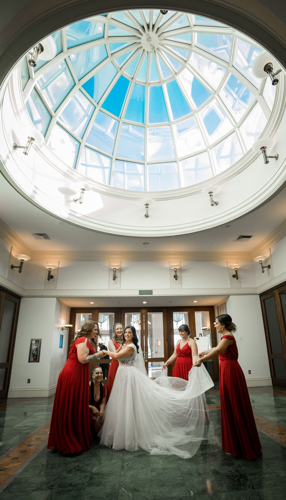 A bridal party helps adjust a wedding dress under an elegant glass dome skylight in a circular rotunda.