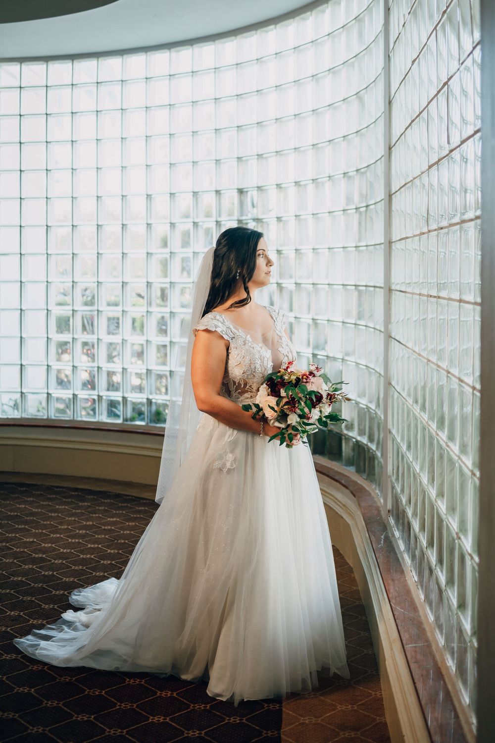 A bride in a flowing white tulle gown holds a pink and white bouquet while standing by a curved glass block wall.