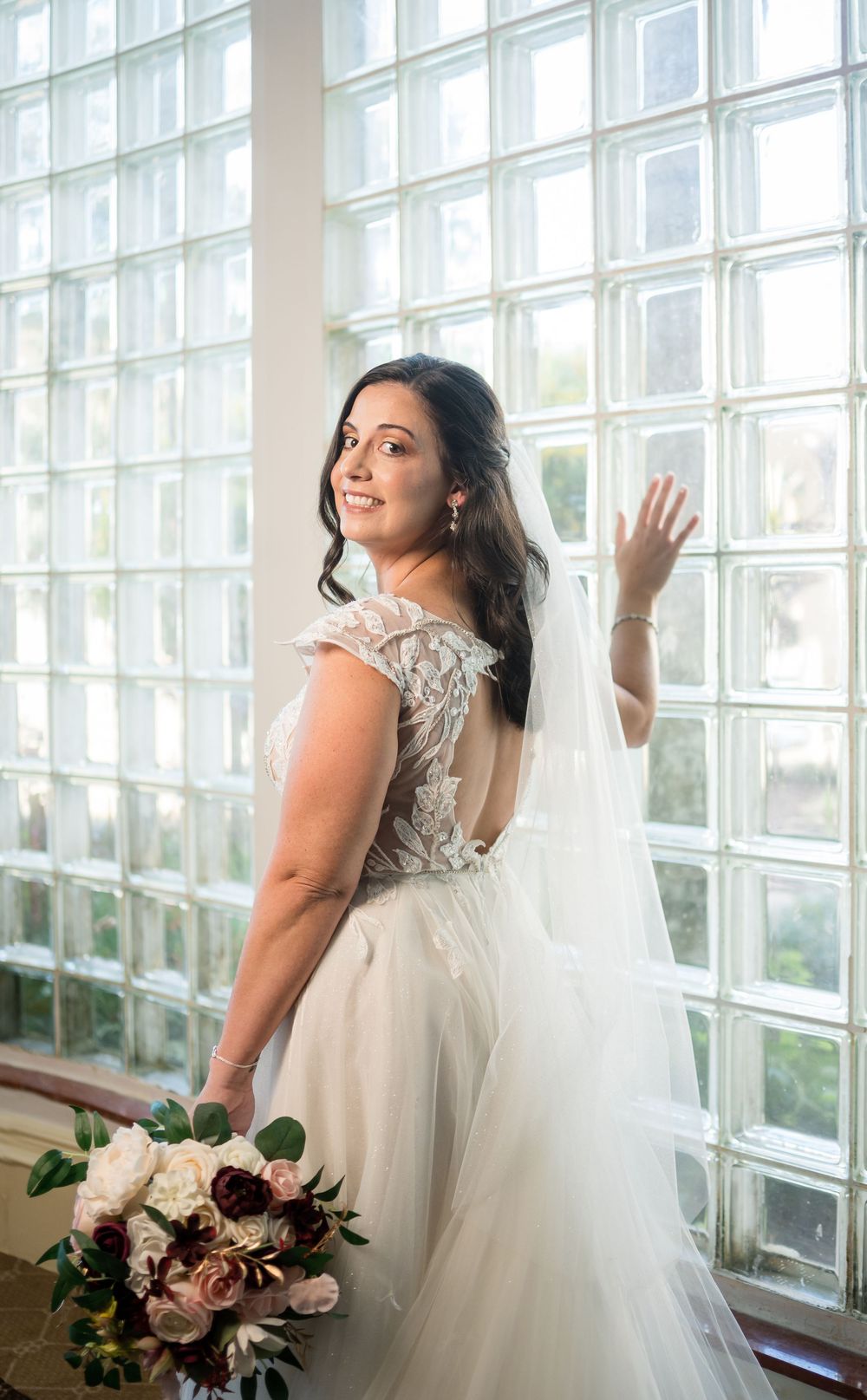 A bride in a lace-back wedding gown with long veil poses by glass block window while holding a pink and cream bouquet.