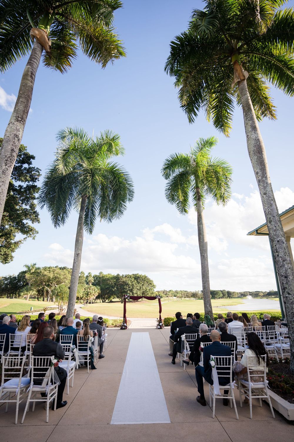 Outdoor wedding ceremony setup with tall palm trees and white chairs along a white aisle runner.