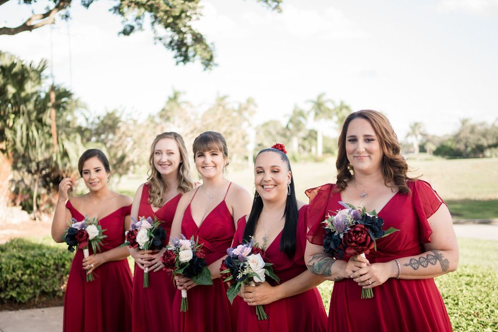 Bridesmaids in vibrant red dresses stand together outdoors holding bouquets on a sunny day at a wedding venue.
