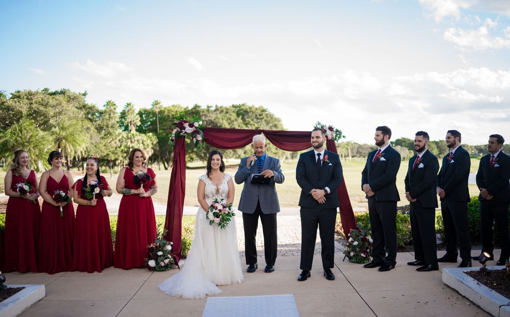 Wedding ceremony outdoors with bridesmaids in burgundy dresses and groomsmen in dark suits arranged around officiant.