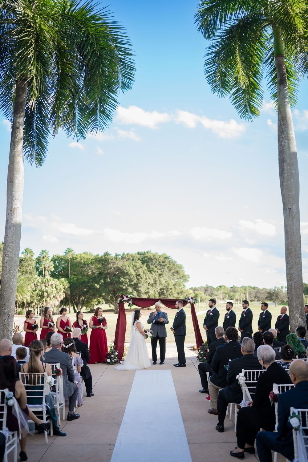 Outdoor wedding ceremony with palm trees framing a white aisle runner and guests seated under bright blue Florida sky.