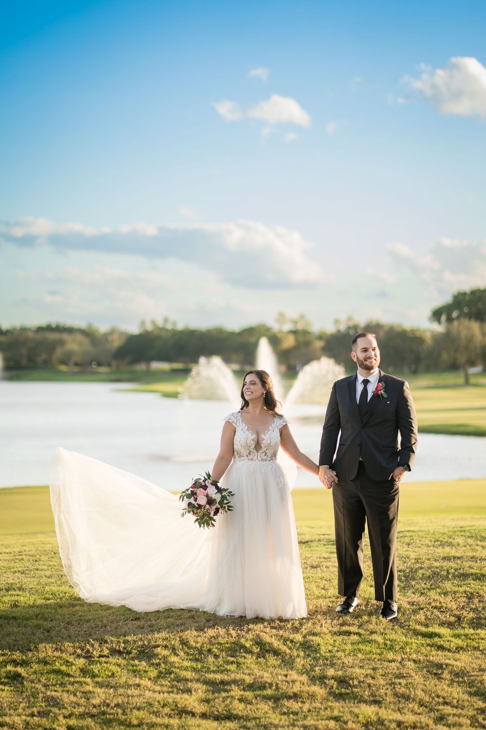 Newlyweds walk together on golf course with water fountain and blue sky in background.