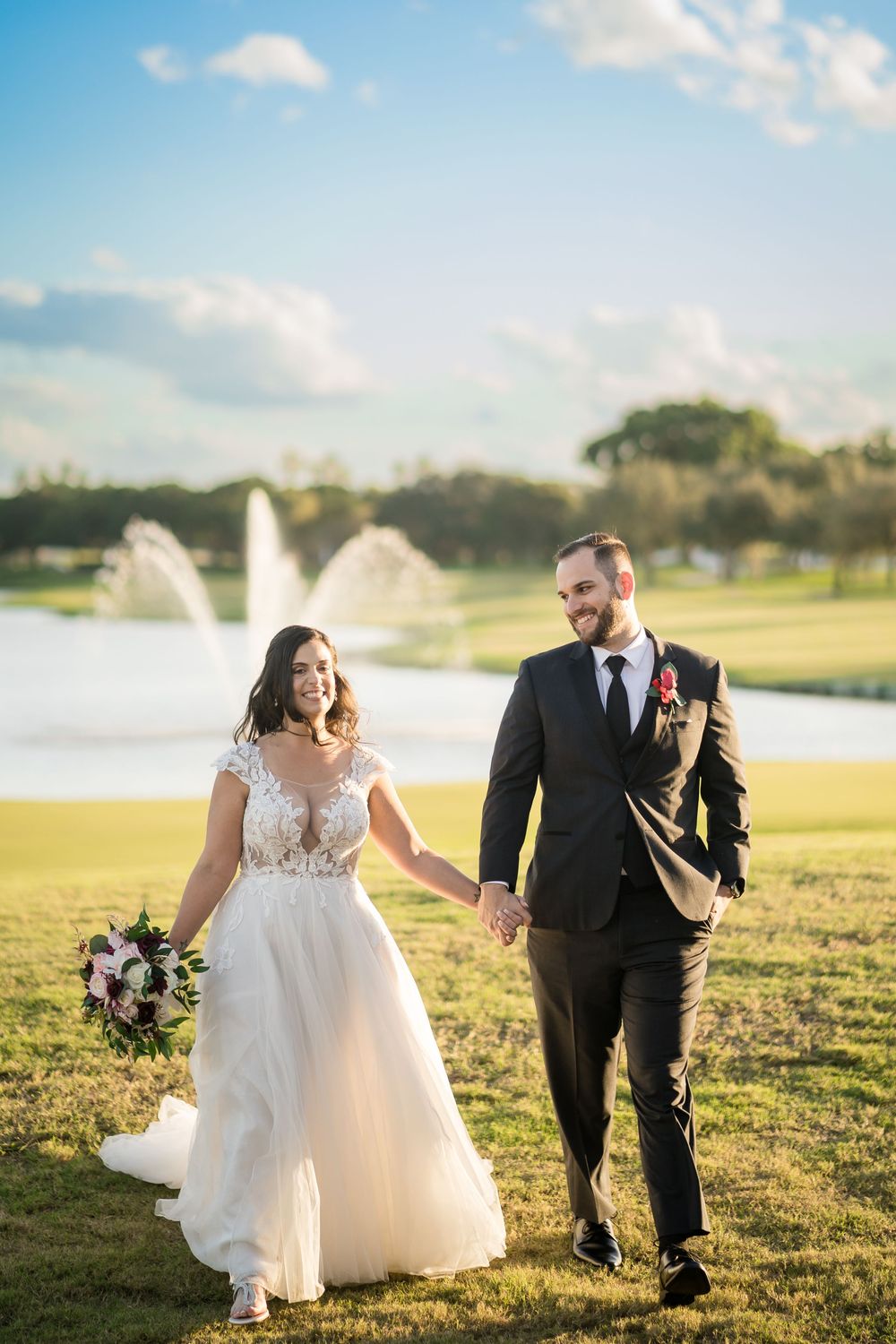 A couple in wedding attire walks hand in hand across a sunny golf course with fountains and water features in the background.