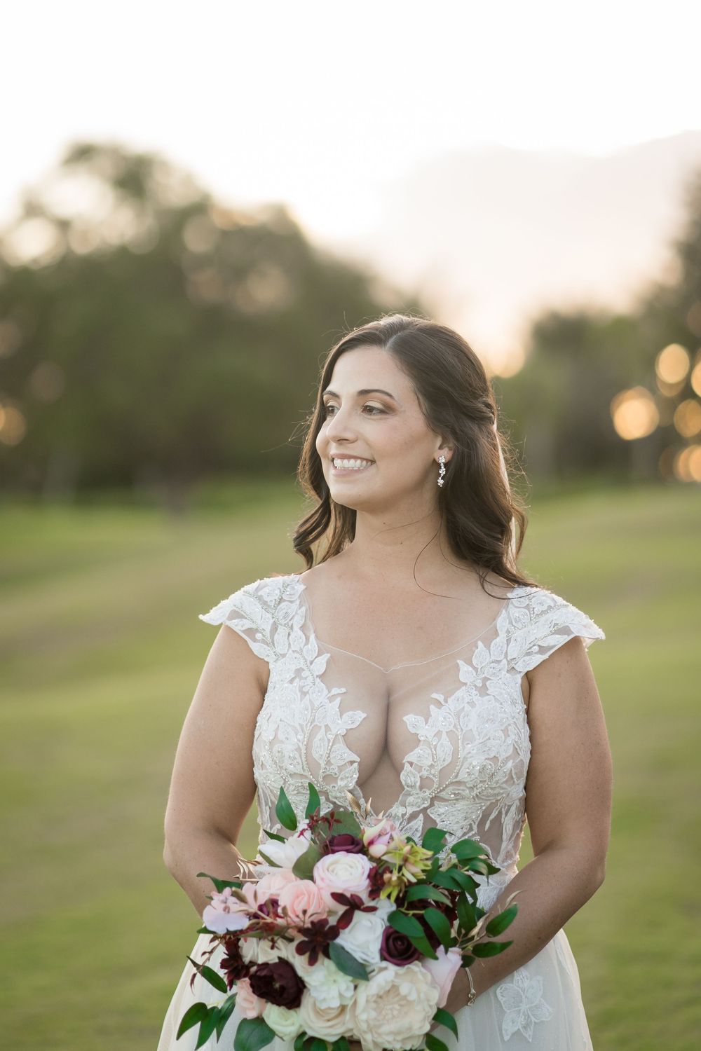 A bride holding a bouquet with burgundy and blush flowers poses in a lace gown during golden hour.
