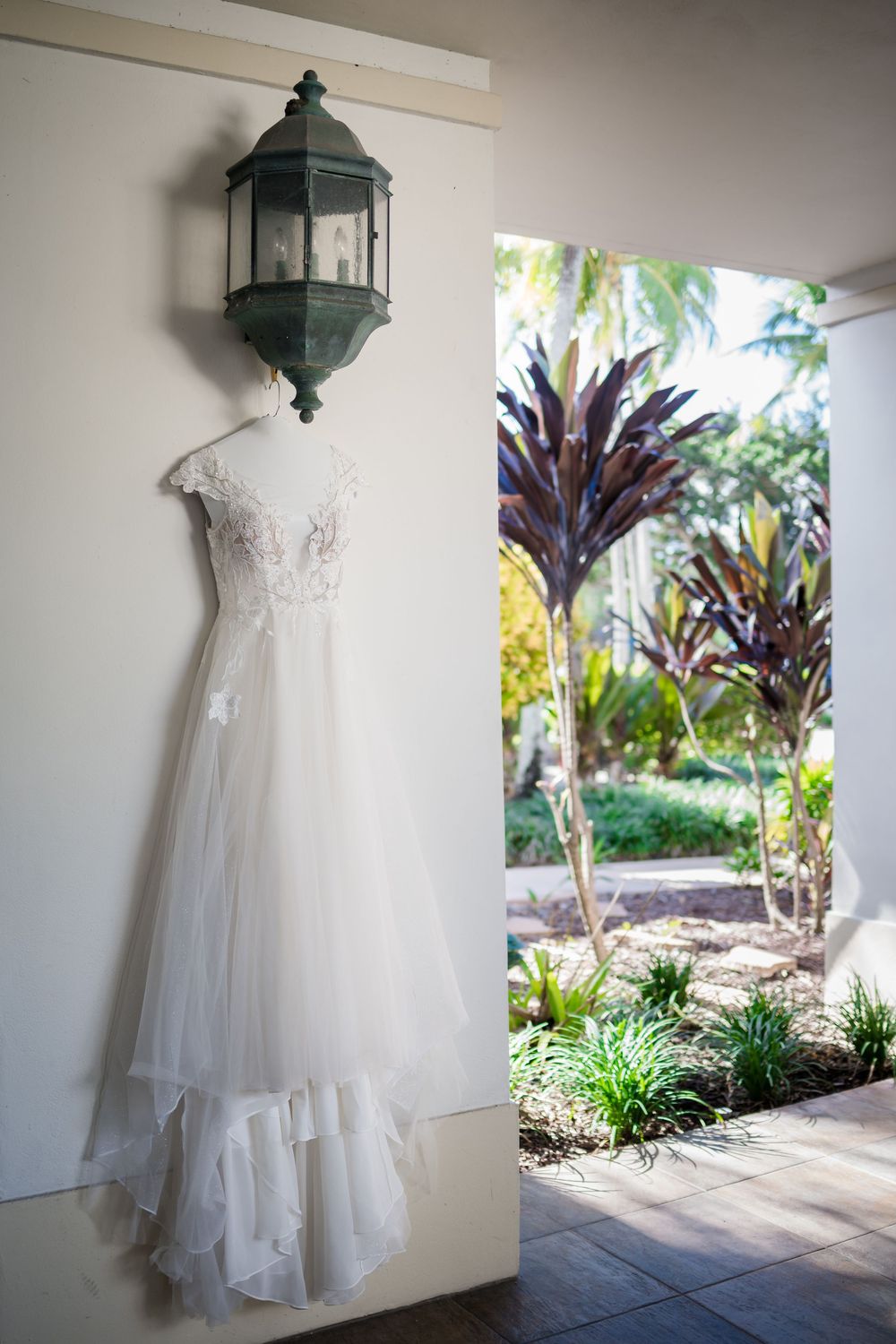 White lace wedding dress hanging beneath vintage lantern on porch with tropical garden view.
