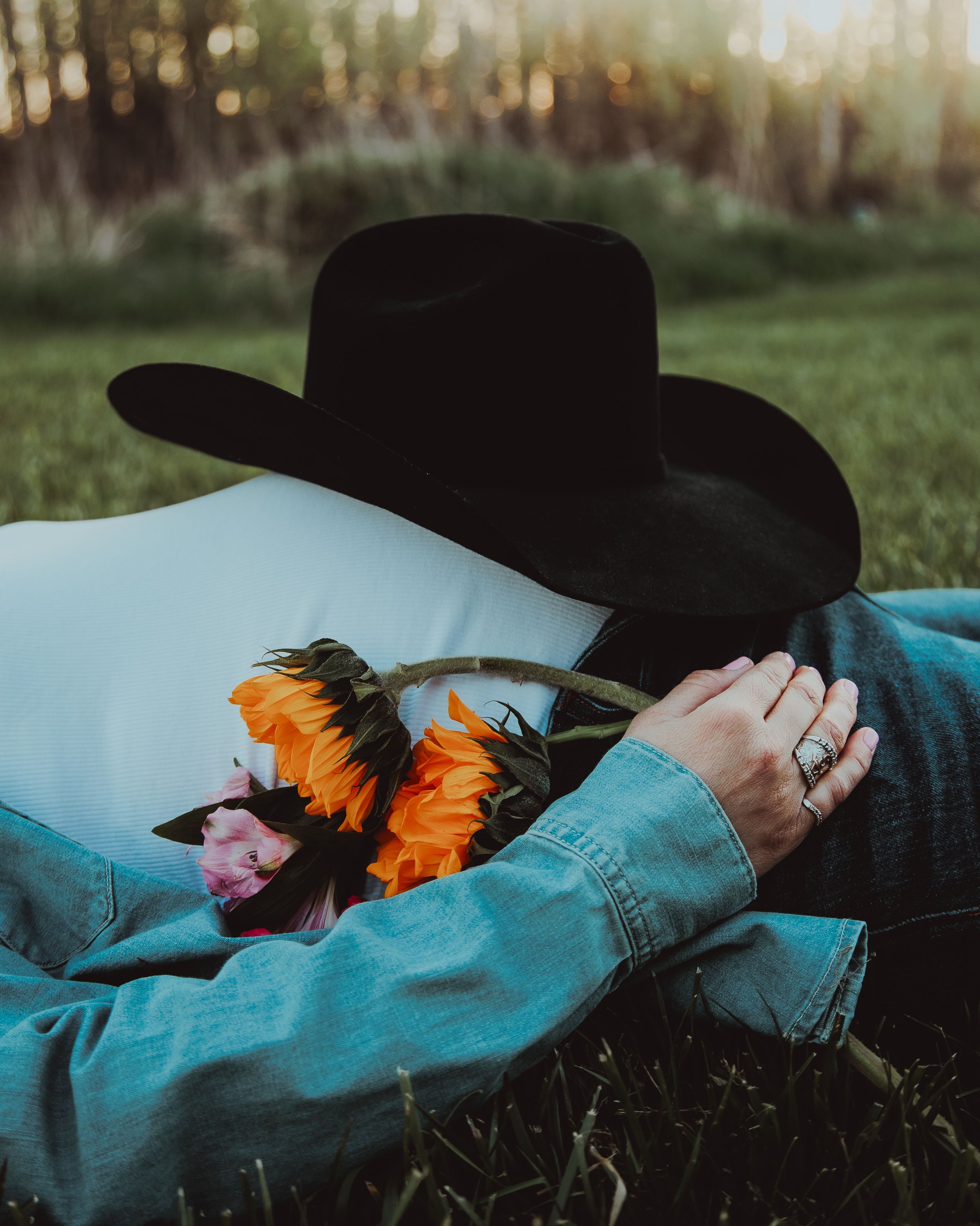 Someone in a denim jacket lying on grass with a black cowboy hat and vibrant orange sunflowers at sunset.