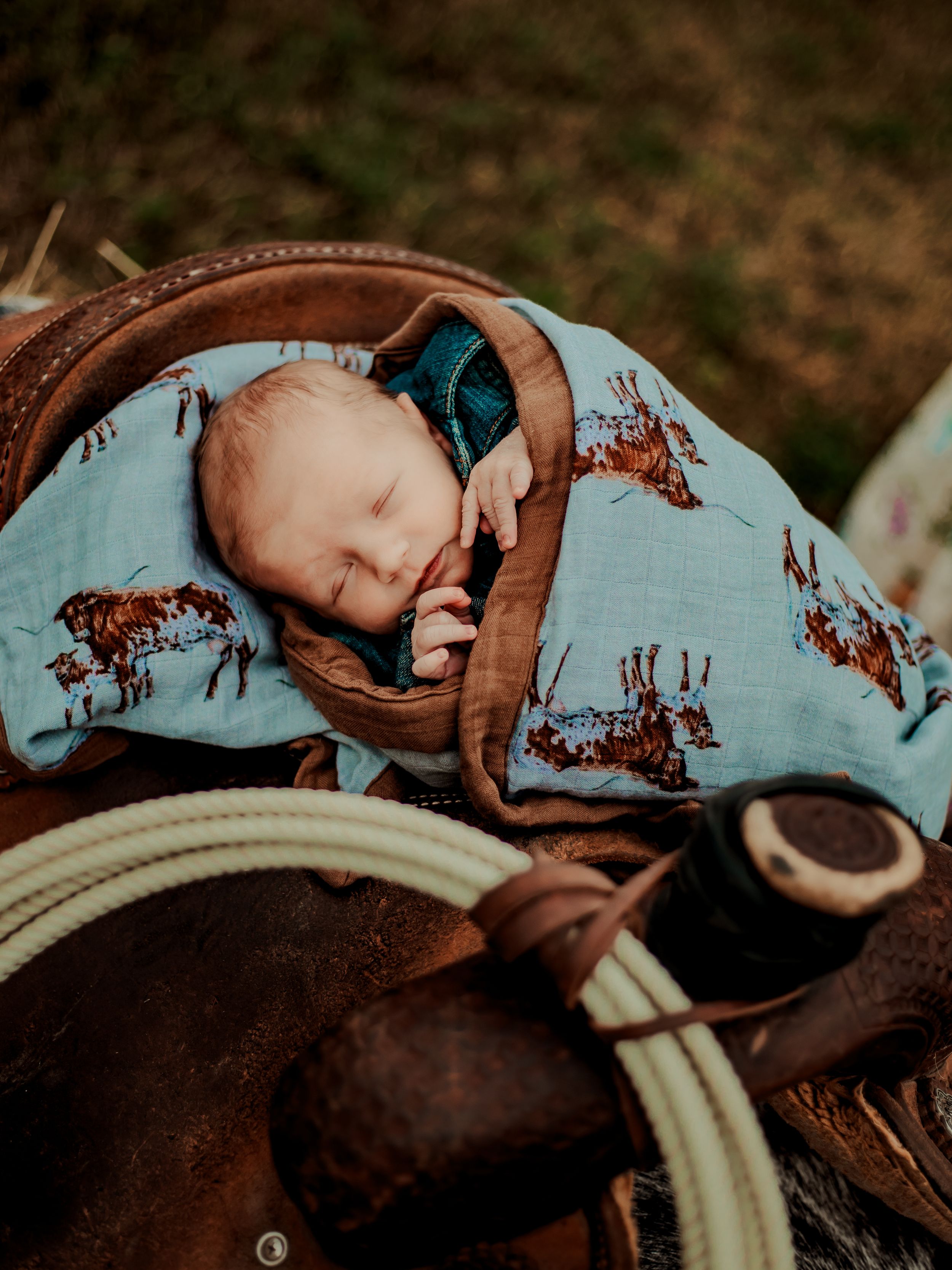 A sleeping infant wrapped in a blue patterned blanket rests on western leather tack and rope.