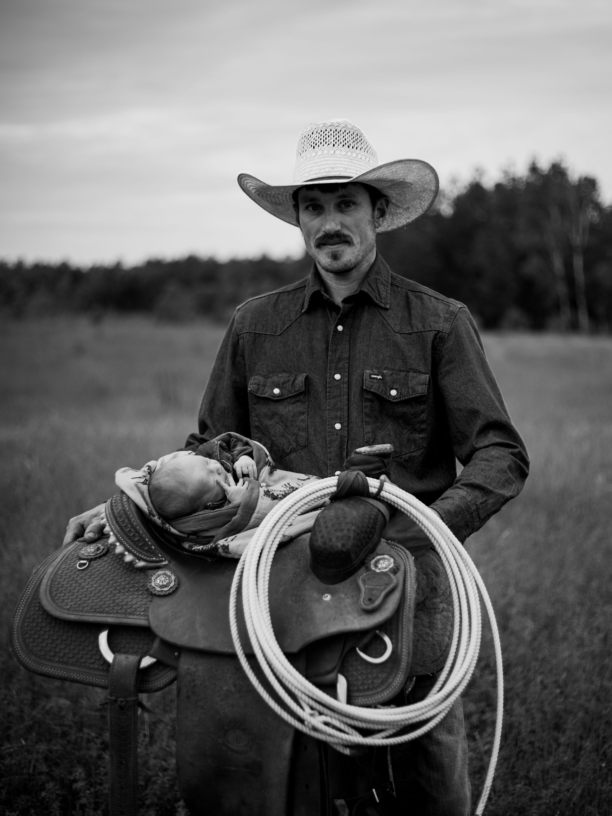 A cowboy in western attire stands with a saddle and lasso rope in a rustic field setting.