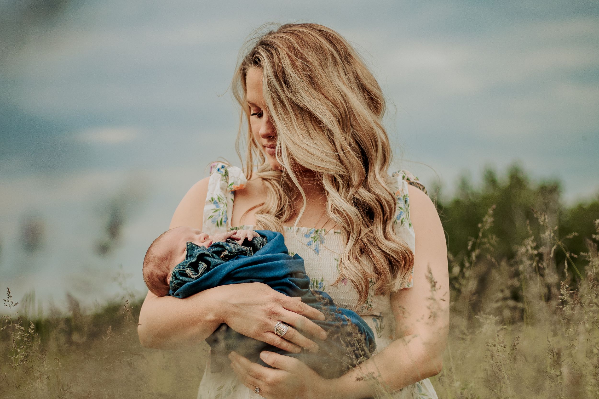 A woman in a blue dress stands in a field of tall grass against a cloudy sky with wavy blonde hair.