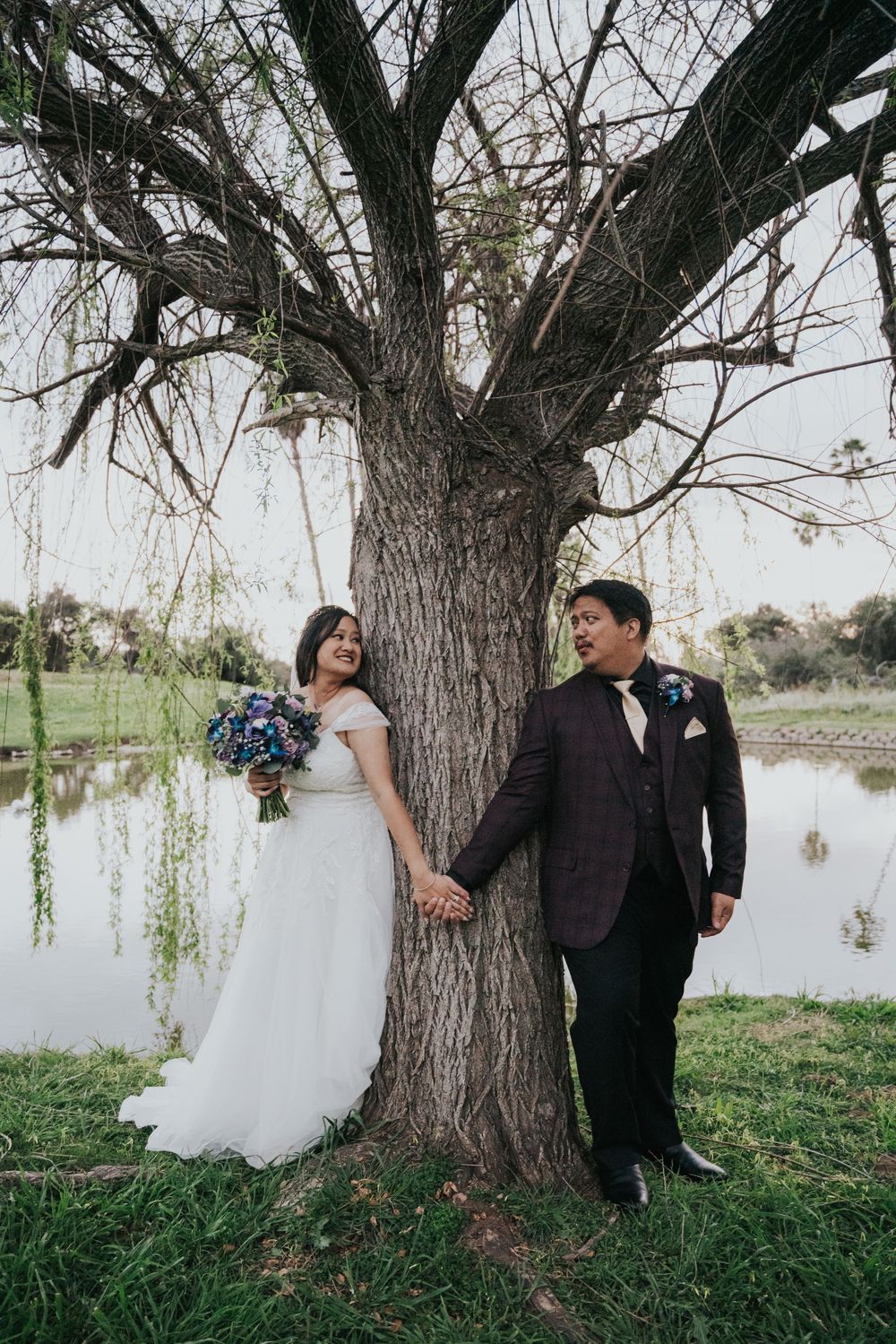 Wedding couple stands by large tree trunk near water, bride in white dress holding blue bouquet and groom in burgundy suit.