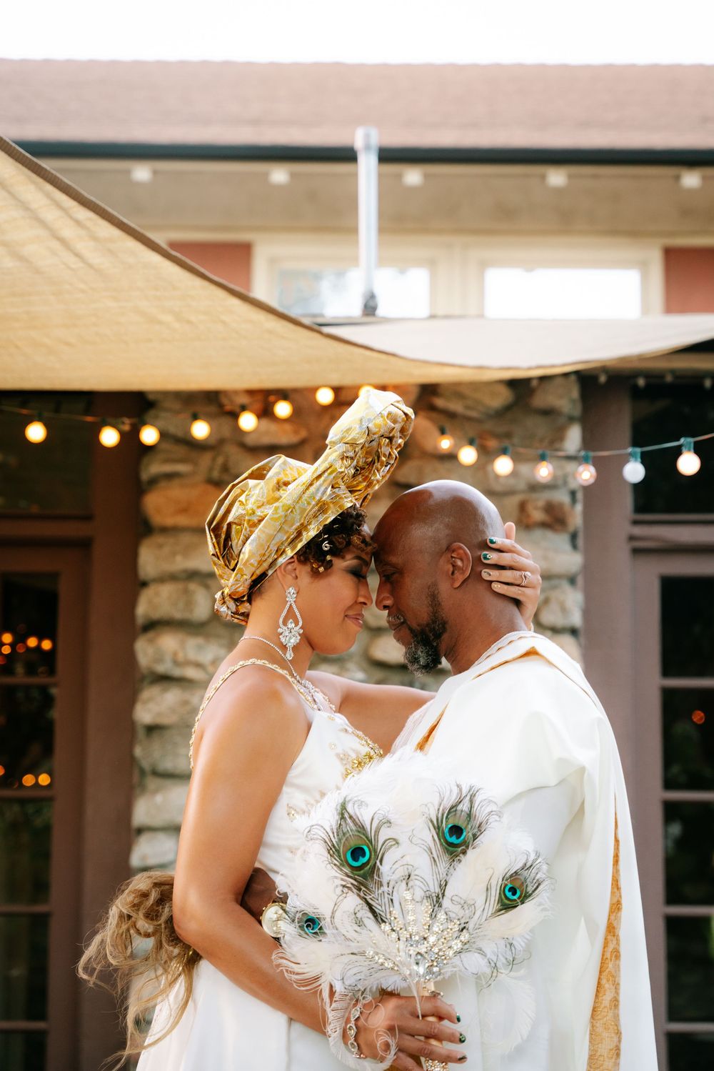 Romantic wedding couple shares an intimate moment under string lights with a peacock feather bouquet and gold accents.