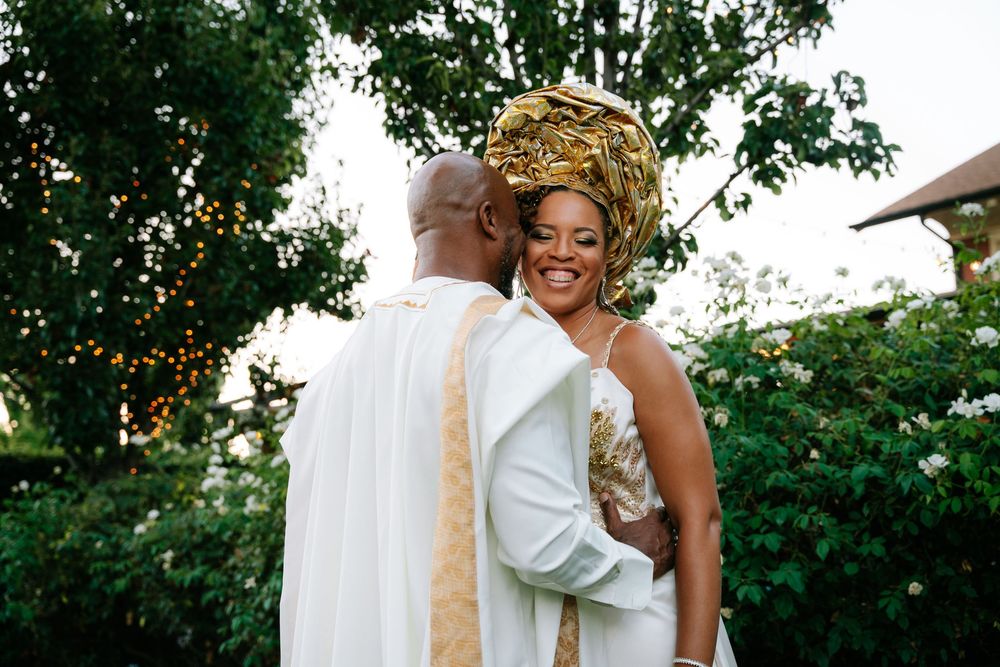 A beaming couple in white and gold traditional African wedding attire embrace outdoors during their celebration.