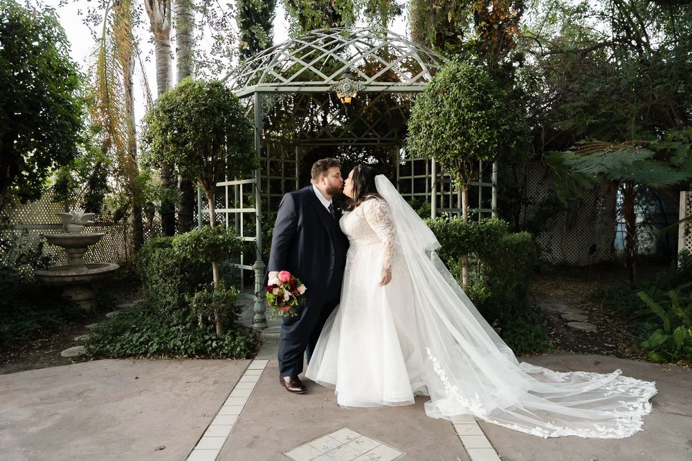 A wedding couple stands together under an elegant outdoor gazebo surrounded by greenery and tall trees.