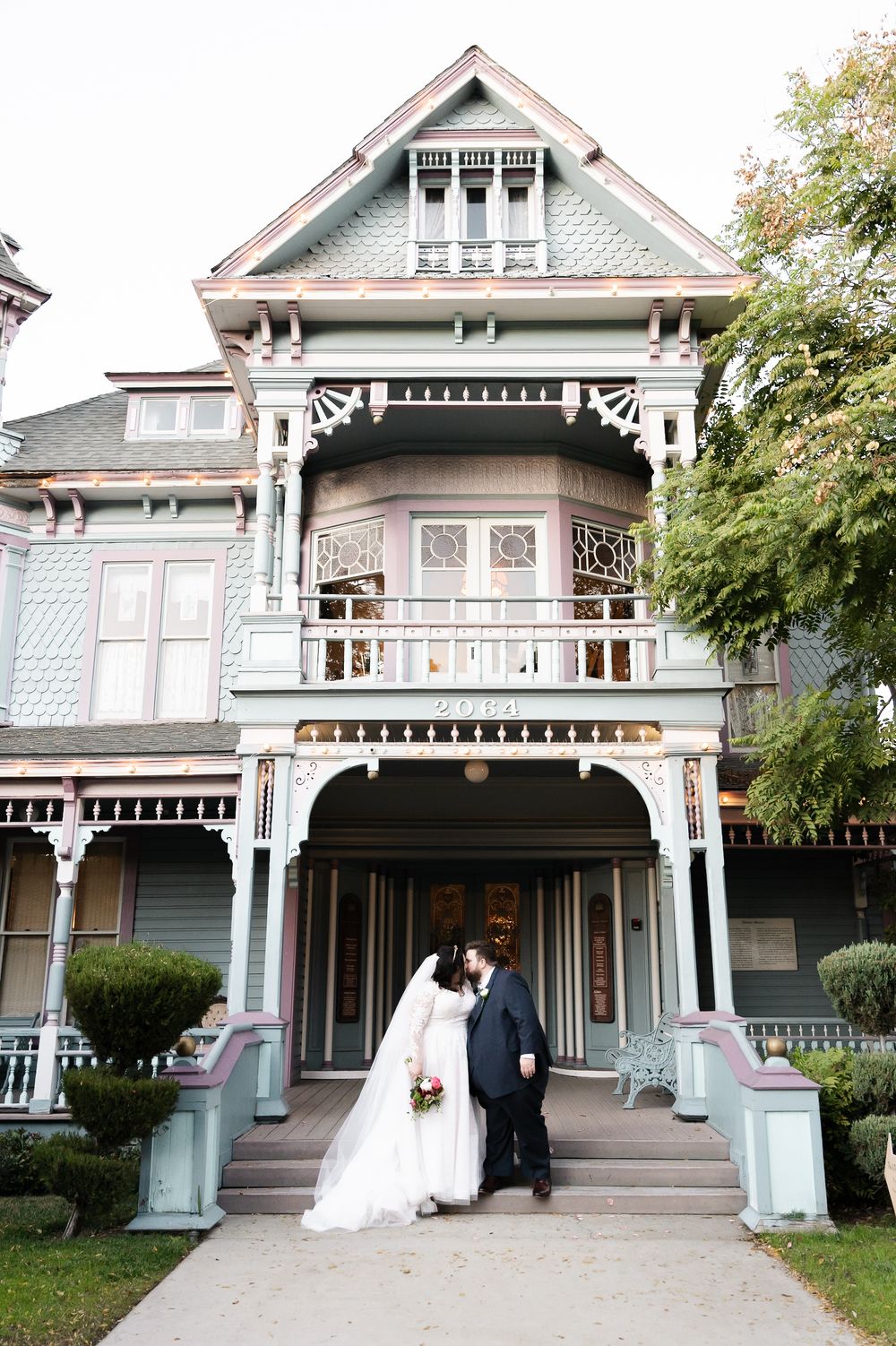 A couple stands on the front steps of an elegant Victorian mansion with ornate trim and pale blue exterior paint.