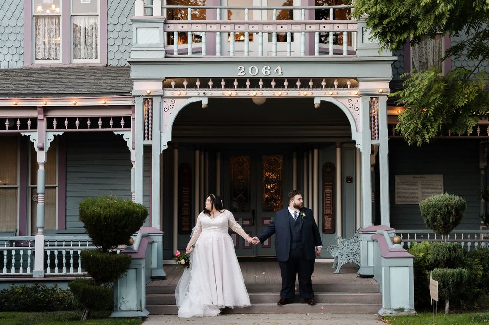 A couple in formal wedding attire stands together on the porch of a Victorian-style house with ornate railings and trim.