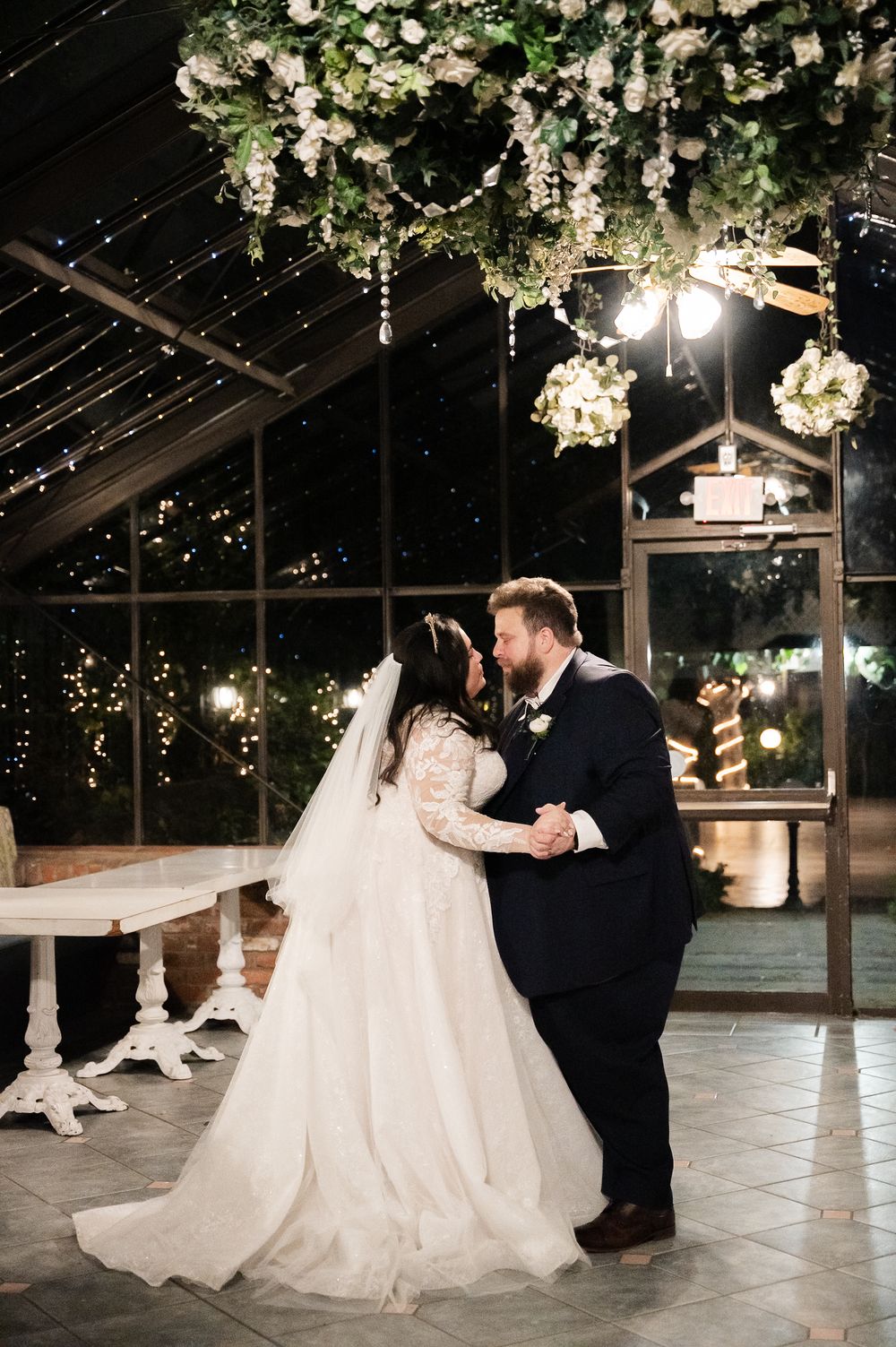 A couple shares their first dance beneath hanging floral arrangements and twinkling lights in an elegant greenhouse venue.