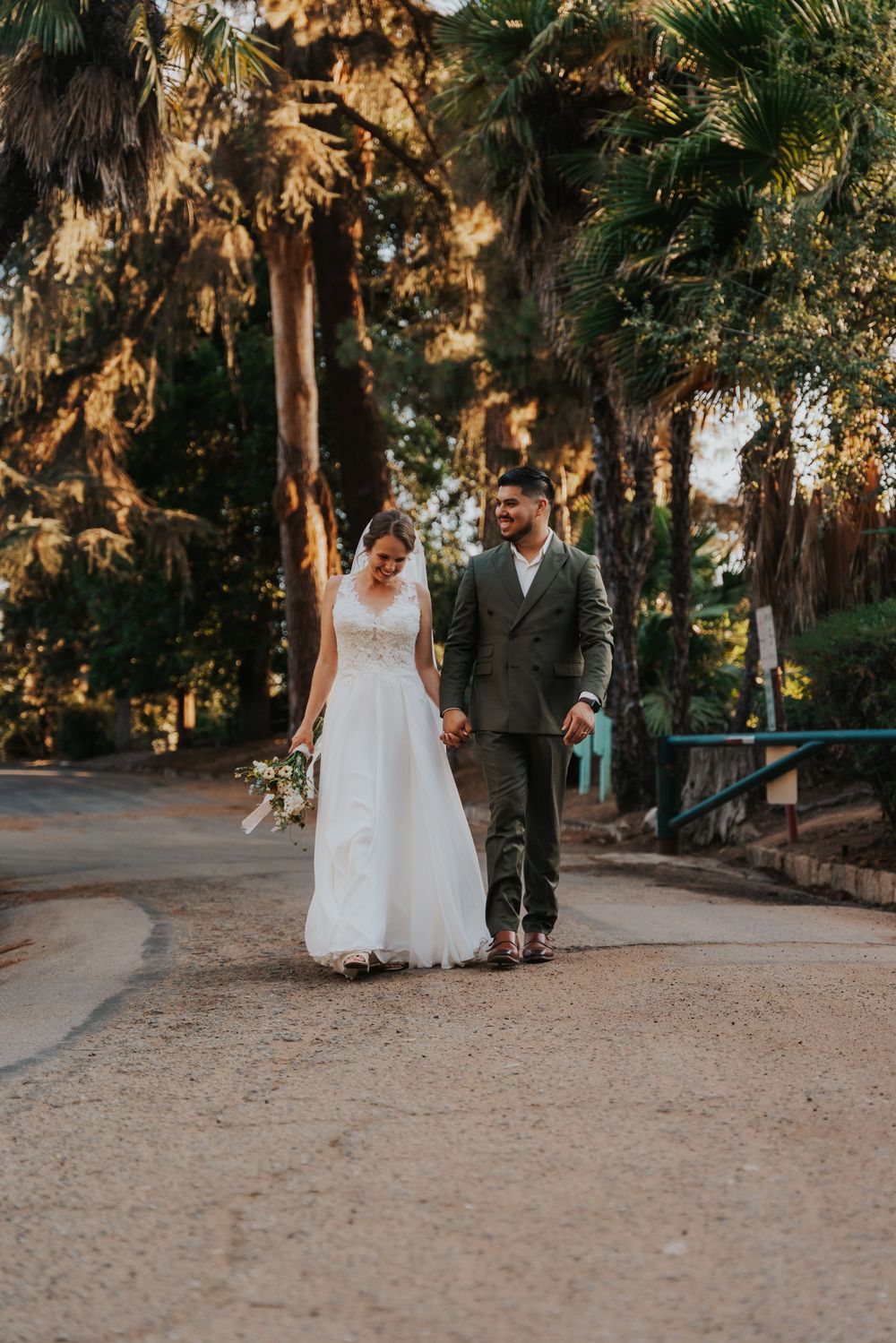A couple in wedding attire walks together on a path surrounded by palm trees and golden sunlight.