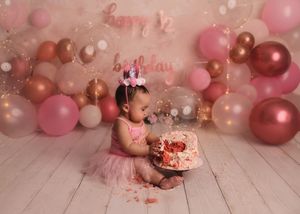 Baby girl in pink tutu smashing first birthday cake surrounded by pink and rose gold balloon backdrop.