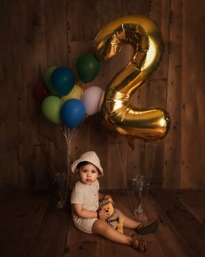 Toddler in white hat sits with teddy bear beside colorful balloons and large gold number 2 balloon for birthday.