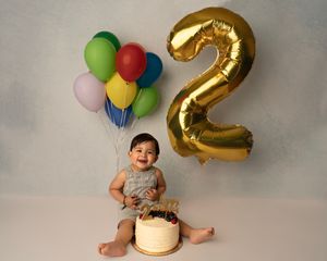 Baby boy smiles at his 2nd birthday cake smash with colorful balloons and a gold number 2 balloon.