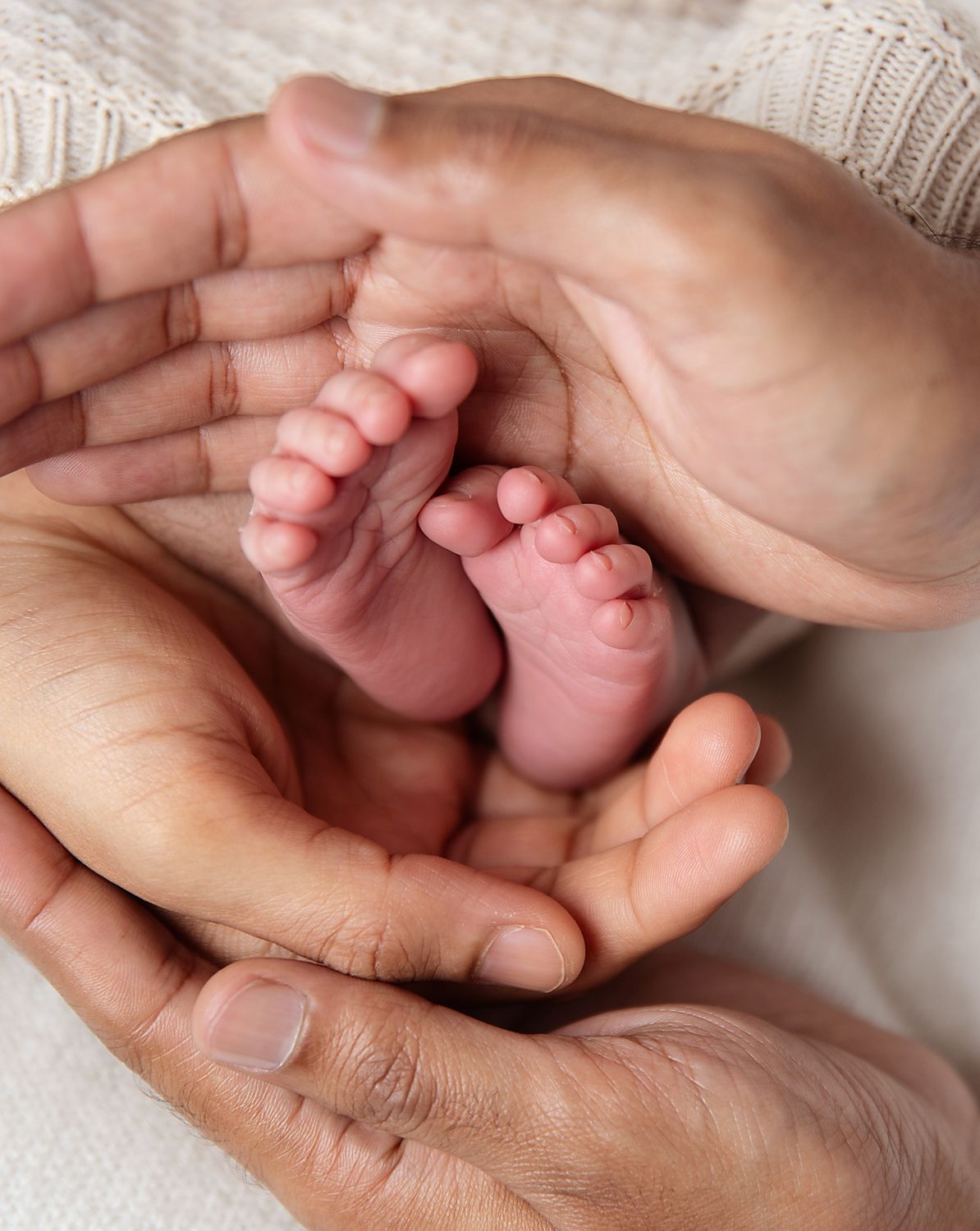 Adult hands gently cradle tiny baby feet against soft white knit fabric in an intimate portrait. During a affordable London baby photoshoot.